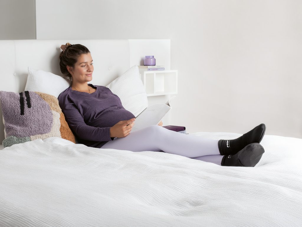Schwangere Frau liegt im Bett und liest ein Buch, hat dabei die Wellness-Kühlsocken an